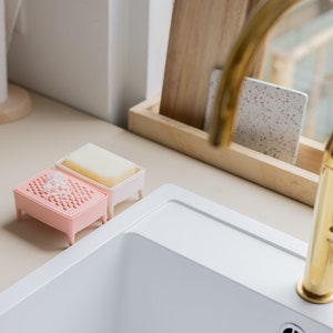 May include: A pink and white soap dish set with a bar of soap and a sponge. The set is on a beige countertop next to a white sink and a gold tap. A wooden tray holds a chopping board and a speckled tile.