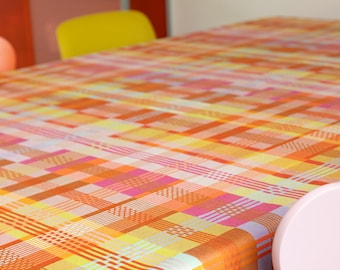 May include: A colorful tablecloth with a plaid pattern in shades of orange, yellow, pink, and white. The fabric is draped over a table, with a portion of a pink chair visible in the foreground. The overall theme is home decor.