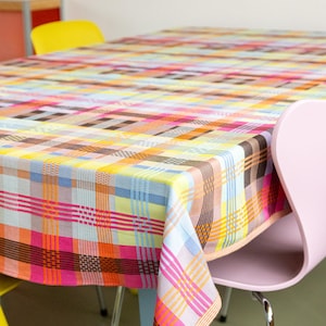 Op de afbeelding: Een kleurrijk geruit tafelkleed in roze, oranje, geel, blauw en bruin bedekt een tafel. Het tafelkleed heeft een rasterpatroon en is gedrapeerd over een tafel met een lichtblauwe basis. Een gele en een roze stoel zijn zichtbaar.