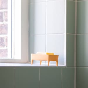 May include: A yellow soap dish with four legs sits on a white countertop in a bathroom. The dish is empty, but there are two bars of soap on top of it.