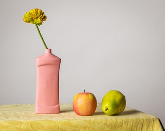 May include: A still life composition featuring a pink vase holding a yellow flower with a green stem. An apple and a pear sit on a yellow tablecloth. The background is a plain white wall.