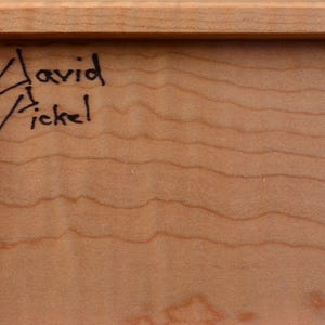 Pode incluir: Close-up de uma moldura de madeira com a assinatura manuscrita "David Fickel" em tinta preta no canto superior esquerdo. A madeira tem uma cor castanho claro com padr&otilde;es de gr&atilde;o vis&iacute;veis.