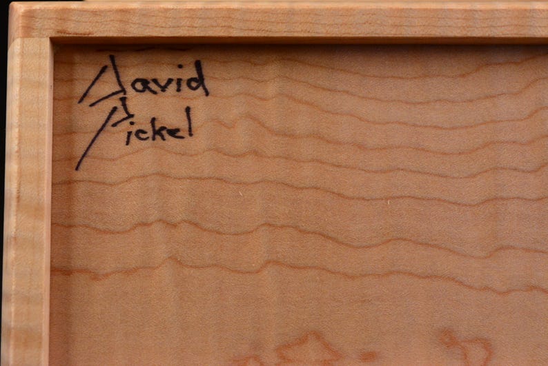 Pode incluir: Close-up de uma moldura de madeira com a assinatura manuscrita "David Fickel" em tinta preta no canto superior esquerdo. A madeira tem uma cor castanho claro com padr&otilde;es de gr&atilde;o vis&iacute;veis.