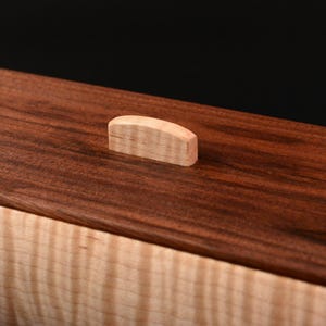 Pode incluir: Uma caixa de madeira com uma parte superior castanha escura e uma base de cor clara. A parte superior apresenta uma pequena pega de madeira de cor clara. O gr&atilde;o da madeira &eacute; vis&iacute;vel, adicionando textura ao design. A caixa est&aacute; sobre um fundo preto.