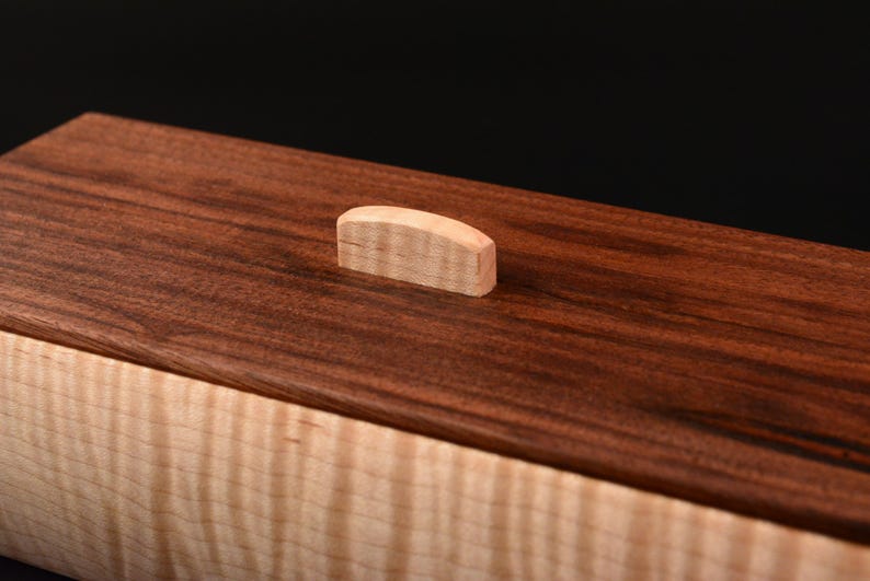 Pode incluir: Uma caixa de madeira com uma parte superior castanha escura e uma base de cor clara. A parte superior apresenta uma pequena pega de madeira de cor clara. O gr&atilde;o da madeira &eacute; vis&iacute;vel, adicionando textura ao design. A caixa est&aacute; sobre um fundo preto.