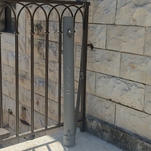 Puede incluir: Un tubo de metal gris con una correa y la palabra hebrea "Jerusalén" escrita en él. El tubo se apoya en una pared de piedra.