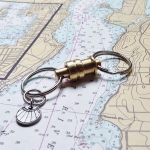 May include: A brass key chain with a compass charm and two silver key rings. The key chain is lying on a nautical chart with numbers and lines.