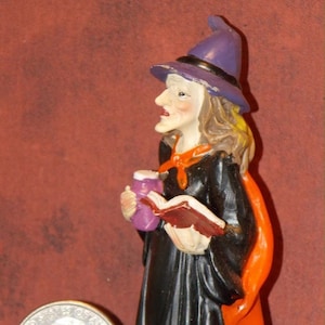 May include: A witch figurine wearing a purple hat, black dress, and orange cape holding a book and a purple potion bottle.