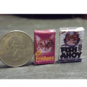 May include: Two miniature boxes of cat food. The box on the left is pink and says "Little Friskies" with a picture of a tabby cat. The box on the right is white and says "Fish Ahoy" with a picture of a tabby cat wearing a sailor hat.