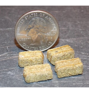 May include: Four miniature hay bales, each about the size of a US quarter, are arranged on a gray surface. The bales are made of dried grass and have a natural brown color.