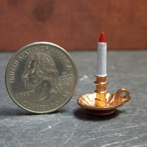 May include: A miniature gold candlestick with a white candle and a red tip. The candlestick is about the size of a US quarter.