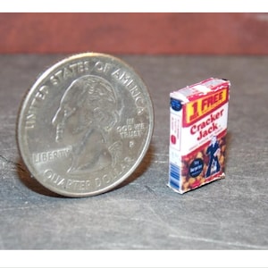May include: Miniature Cracker Jack box with red, white, and blue packaging, next to a U.S. quarter. The box features the text "1 FREE Cracker Jack" and a graphic of a man in a suit. A miniature food item.