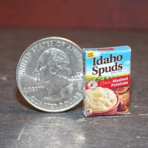 Peut inclure: Une boîte miniature de pommes de terre en purée classiques Idaho Spuds. La boîte est bleue et rouge avec du texte blanc. La boîte est à côté d'un quart de dollar américain pour l'échelle.