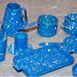 Puede incluir: Un juego de cocina de juguete de esmalte azul y blanco con una jarra, dos ollas, una taza, un plato, una bandeja y un cubo. El juego está hecho de metal y tiene un acabado moteado.