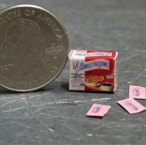 May include: A miniature box of Sweet'N Low sugar substitute with pink packets, next to a US quarter for scale.