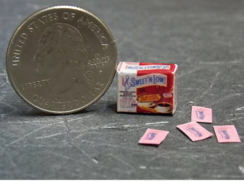 Op de afbeelding: Een miniatuurdoosje Sweet'N Low suikervervanger met roze zakjes, naast een Amerikaanse kwart dollar voor de schaal.