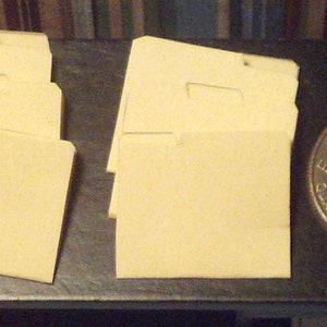 May include: A stack of six cream-colored file folders with a tab on the left side. The folders are arranged in a vertical stack.