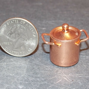 Puede incluir: Una olla pequeña de cobre con tapa y dos asas. La olla es del tamaño de una moneda de euro.