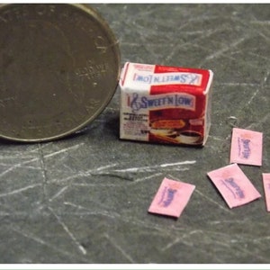 Op de afbeelding: Een miniatuurdoosje met Sweet'N Low suikervervanger, samen met vier miniatuurzakjes Sweet'N Low. Het doosje is rood en wit met het Sweet'N Low-logo. De zakjes zijn roze met het Sweet'N Low-logo.