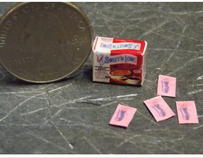 Op de afbeelding: Een miniatuurdoosje met Sweet'N Low suikervervanger, samen met vier miniatuurzakjes Sweet'N Low. Het doosje is rood en wit met het Sweet'N Low-logo. De zakjes zijn roze met het Sweet'N Low-logo.