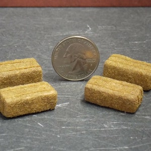 May include: Four miniature hay bales, each about the size of a US quarter, are arranged on a gray surface. The bales are made of a brown, fibrous material and have a slightly rough texture.
