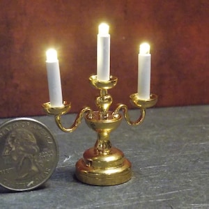 May include: A gold candelabra with three white candles. The candles are lit and the candelabra is sitting on a gray surface.
