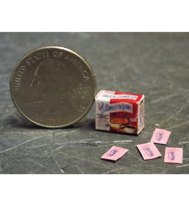 Op de afbeelding: Een miniatuurdoosje met Sweet'N Low suikervervanger, naast een Amerikaanse kwartje, en vier roze Sweet'N Low-zakjes.