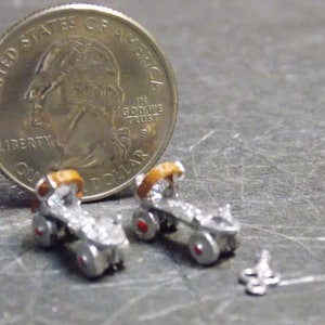 May include: A pair of miniature silver roller skates with brown leather straps and red wheels. The skates are next to a US quarter dollar coin for scale.