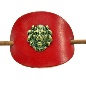 Könnte beinhalten: Eine rote Lederhaarklammer mit einem Holzstift und einem Löwenkopfdesign in der Mitte.