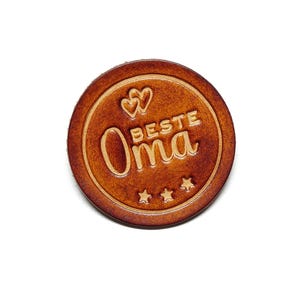 Könnte beinhalten: Ein brauner Lederkreis mit dem Text "BESTE Oma" in hellbrauner Farbe. Um den Text herum befinden sich zwei Herzen und drei Sterne.