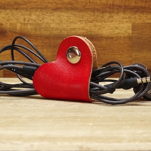Könnte beinhalten: Roter Leder-Kopfhörerhalter in Herzform mit einem schwarzen Kabel, das darum gewickelt ist. Das Kabel hat einen schwarzen und silbernen Kopfhöreranschluss mit dem Text "AKG" darauf.