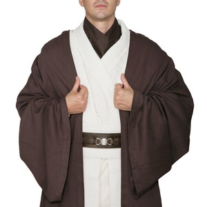 May include: A brown Jedi robe costume with a white tunic and brown belt. The costume features wide sleeves and a high collar. The belt has a silver buckle. The person is wearing the costume and looking at the camera.