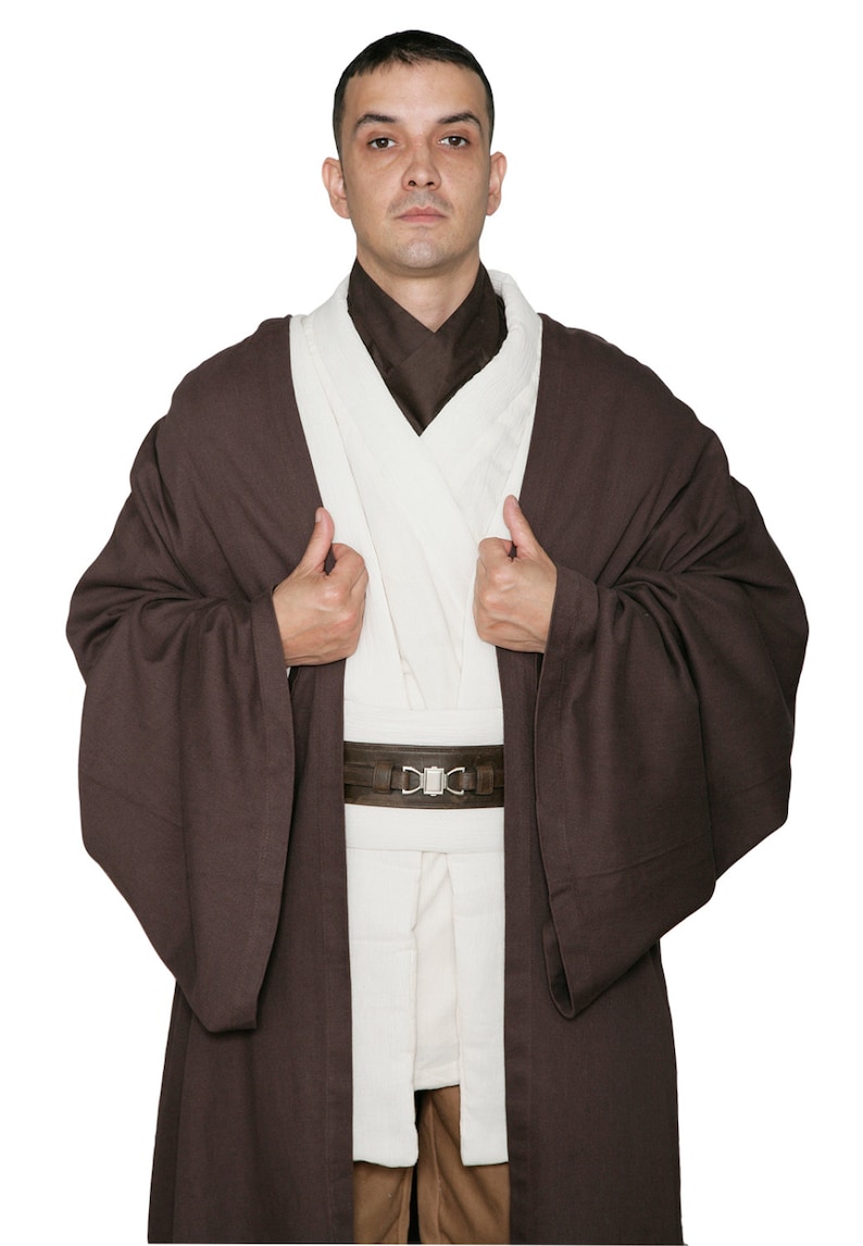May include: A brown Jedi robe costume with a white tunic and brown belt. The costume features wide sleeves and a high collar. The belt has a silver buckle. The person is wearing the costume and looking at the camera.
