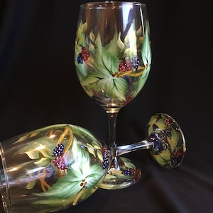 May include: Two clear wine glasses with hand-painted designs. The glasses feature green leaves, red and blue berries, and yellow accents. One glass is upright, and the other is lying on its side.