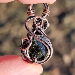 May include: A handcrafted pendant featuring a dark green stone encased in copper wire. The wire is intricately wrapped around the stone, forming a decorative design. The pendant is held up by a finger.