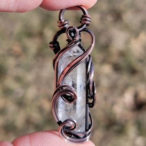 May include: A clear quartz crystal pendant wrapped in copper wire. The wire is intricately coiled around the crystal, forming decorative swirls and a loop for a chain. The pendant is held between fingers against a blurred background.