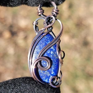 May include: A handcrafted pendant featuring a vibrant blue gemstone, intricately wrapped with copper wire. The wirework forms decorative swirls and frames the stone, creating a unique and artistic piece of jewelry.
