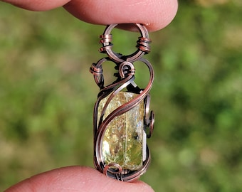 May include: A pendant featuring a translucent, pale yellow, rectangular gemstone encased in dark, oxidized copper wire wrapping. The wire forms intricate, swirling patterns around the stone and includes a circular loop at the top for hanging.