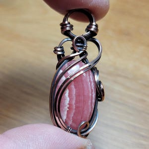 May include: A handcrafted pendant featuring a pink and white striped gemstone, intricately wrapped in copper and silver-toned wire. The pendant has a loop at the top for a chain and a unique, artistic design. The gemstone is approximately 1.5 inches long.