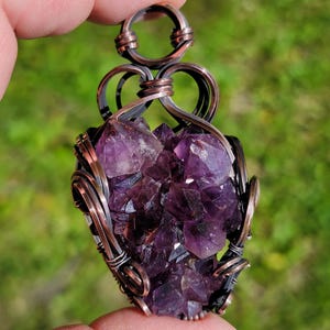 May include: A handcrafted pendant featuring a cluster of deep purple amethyst crystals. The crystals are encased in a copper wire wrap with intricate looping and swirling designs. The pendant has a bail for hanging and is held against a blurred green background.