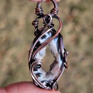 May include: A handcrafted pendant featuring a geode stone with a black, white, and gray pattern. The stone is encased in copper wire with a loop for a necklace. The geode has a crystal-filled center.