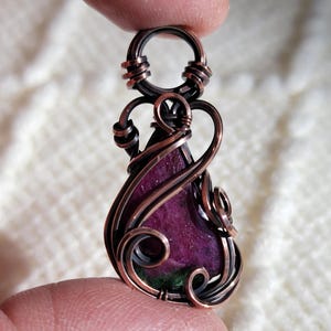 May include: A handcrafted pendant featuring a teardrop-shaped, deep magenta gemstone with green inclusions, encased in intricately woven copper wire. The wirework forms decorative swirls and loops, creating a unique, artistic design. The pendant is held by a finger.