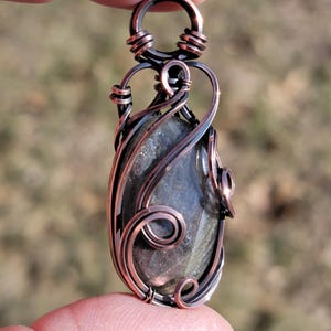 May include: A handcrafted copper wire-wrapped pendant featuring a polished, oval-shaped stone with gray and green hues. The wirework includes intricate swirls and loops, creating a unique, artistic design. The pendant is held between fingers.