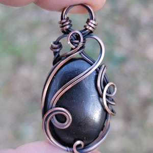 May include: A handcrafted pendant featuring a polished, oval-shaped black stone encased in copper wire. The wire is intricately wrapped and shaped into decorative swirls and loops, creating a unique and artistic design. The pendant is held by a finger.