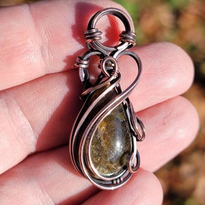 May include: A handcrafted copper wire pendant with a teardrop-shaped, olive-green gemstone. The pendant features intricate wire wrapping and a loop for a chain. The gemstone has a textured appearance, and the copper wire has a dark, aged finish.