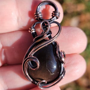 May include: A handcrafted pendant featuring a polished, dark gemstone encased in intricate copper wire. The wirework includes swirling patterns and a bail for a chain. The gemstone has a reflective surface, and the pendant is held in a hand.