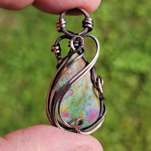 Puede incluir: Un colgante en forma de lágrima con una piedra multicolor que presenta inclusiones verdes, amarillas y rosas, engastada en un marco de alambre de cobre oscuro. El diseño de alambre incluye espirales intrincadas y una argolla circular.