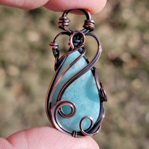 May include: A handcrafted pendant featuring a teardrop-shaped, light blue gemstone. The stone is encased in intricate copper wire, with decorative swirls and loops. The pendant is suspended from a copper wire bail.