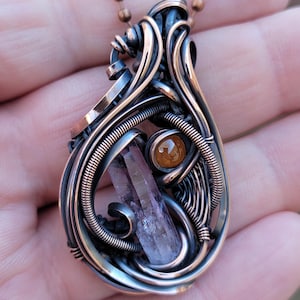 May include: A handcrafted pendant with a wire-wrapped design. The pendant features a teardrop shape with a central amethyst crystal and a smaller orange-brown stone. The metalwork is a mix of silver and copper tones, suspended from a copper-colored chain.