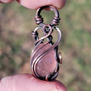 May include: A handcrafted pendant featuring a pink gemstone wrapped in copper wire. The wire is intricately woven around the stone, creating a decorative design. The pendant is held up by a loop at the top, ready to be worn as a necklace.